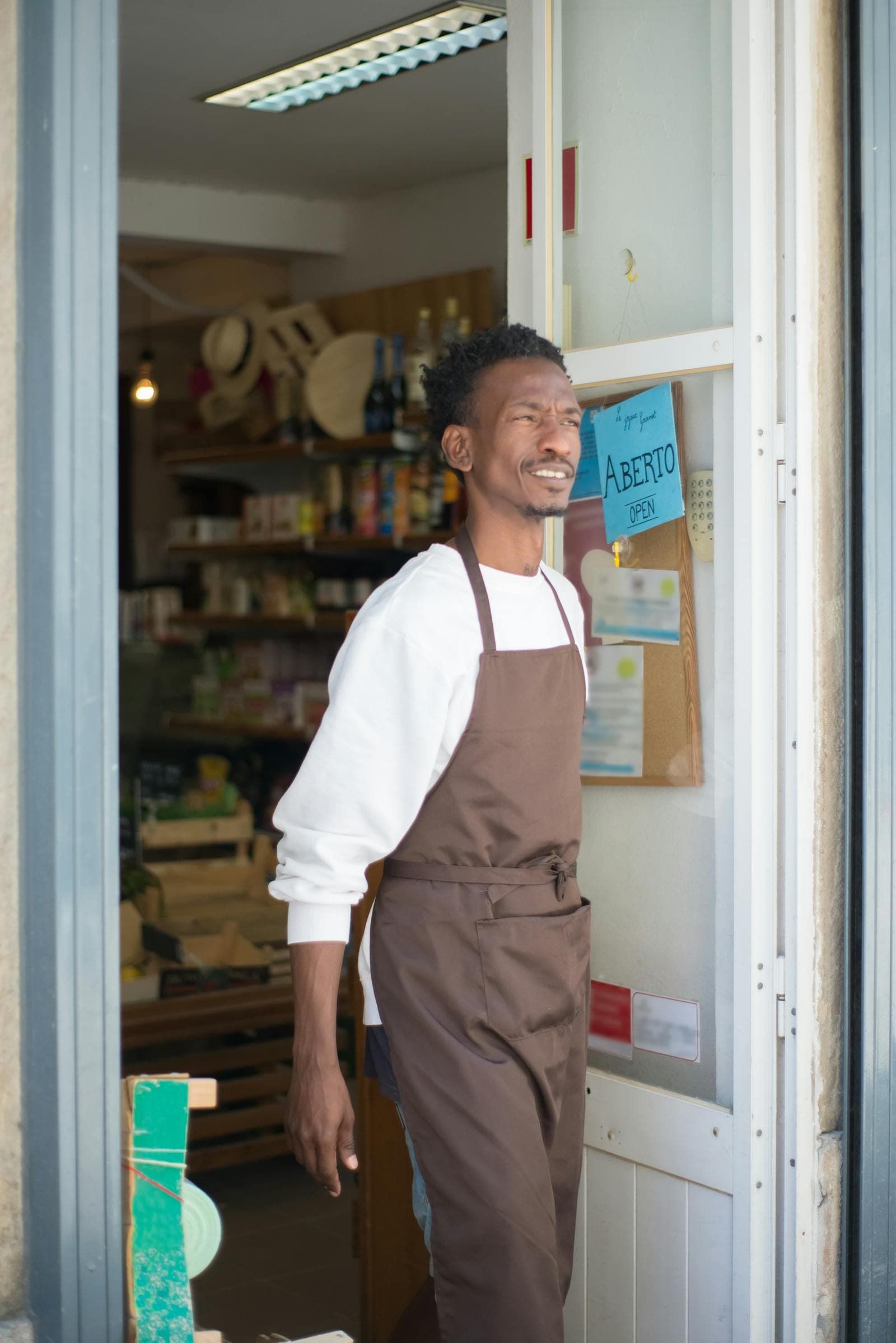 A smiling shop owner in an apron standing in the open doorway of his store.